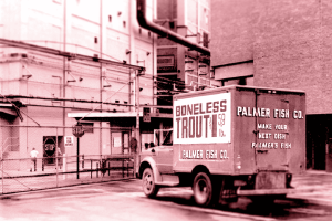 A red-toned picture of an old box truck draped parked in a back building lot and advertising boneless trout.